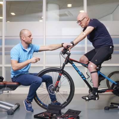 Rembrandt Fysiotherapie & Revalidatie - Zadelpijn en bikefitting Fysiotherapeut Roel in zijn blauwe shirt helpt Evert op de fiets door middel van bikefitting om van zijn zadelpijn af te komen.