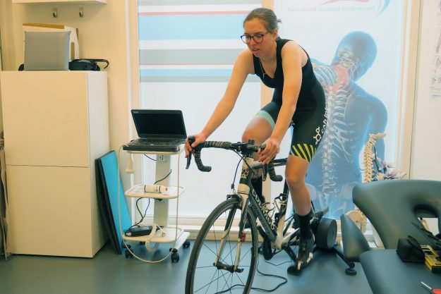 Meisje met bril zit in een wielrennerpak op een racefiets binnen de fysiotherapiepraktijk voor bikefitting en is aan het fietsen.
