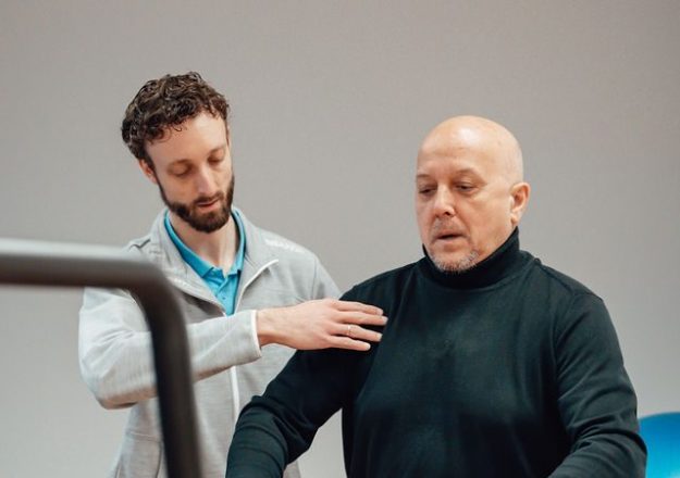 Theo in zijn zwarte shirt wordt ondersteund door fysiotherapeut Bas van Silfhout in een grijze hoodie tijdens oefeningen voor sporten en bewegen met parkinson.