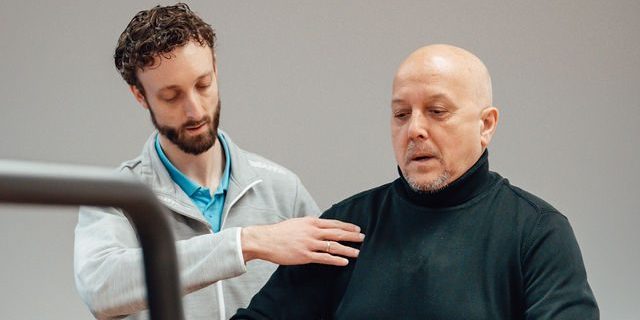Rembrandt fysiotherapie & revalidatie – sporten en bewegen met parkinson Theo in zijn zwarte shirt wordt ondersteund door fysiotherapeut Bas van Silfhout in een grijze hoodie tijdens oefeningen voor sporten en bewegen met parkinson.