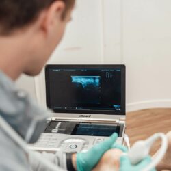 Fysiotherapeut in een grijze polo laat door middel van echografie de hiel pees van een patiënt zien bij 65 plus.