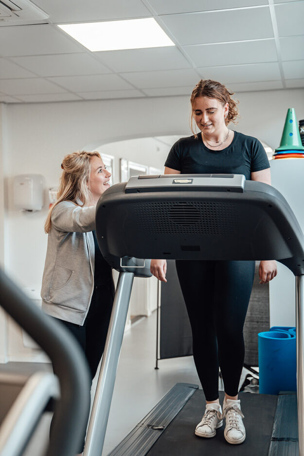 vrouw in een zwart sportshirt werkt aan haar warming-up in de behandelruimte op een loopband. Er staat een fysiotherapeut naast.