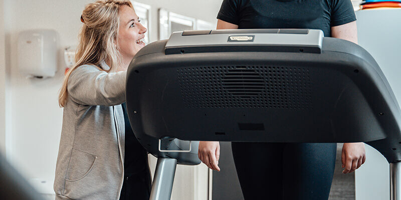 cbfd3e1b-4699-4ef0-b2da-94aa31c952ba (1) vrouw in een zwart sportshirt werkt aan haar warming-up in de behandelruimte op een loopband. Er staat een fysiotherapeut naast.
