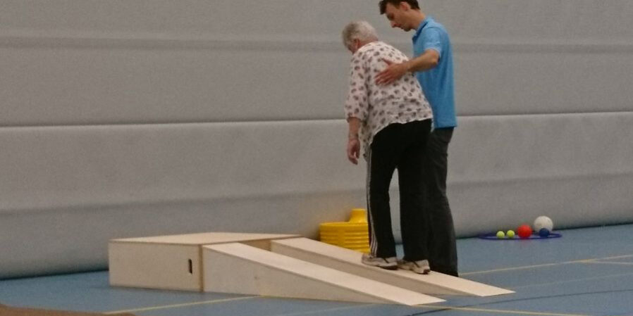 valpreventie rembrandt Vrouw doet aan valpreventie en wordt geholpen door een fysiotherapeut in een blauw shirt in de gymzaal.