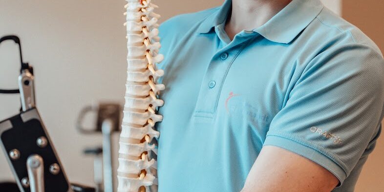Aspecifieke rugklachten Een fysiotherapeut met een lichtblauw t-shirt aan staat in een oefenzaal. Hij houdt de wervelkolom van een skelet vast en kan zo aspecifieke rugklachten uitleggen.