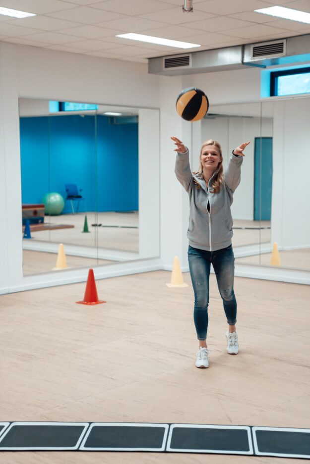 Een vrouw is aan het bewegen. Ze staat in een oefenzaal en ze gooit een bal omhoog.