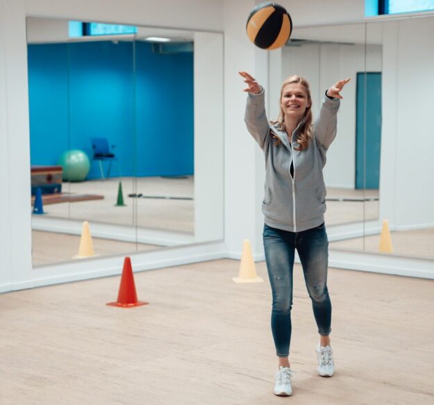 Een vrouw is aan het bewegen. Ze staat in een oefenzaal en ze gooit een bal omhoog.