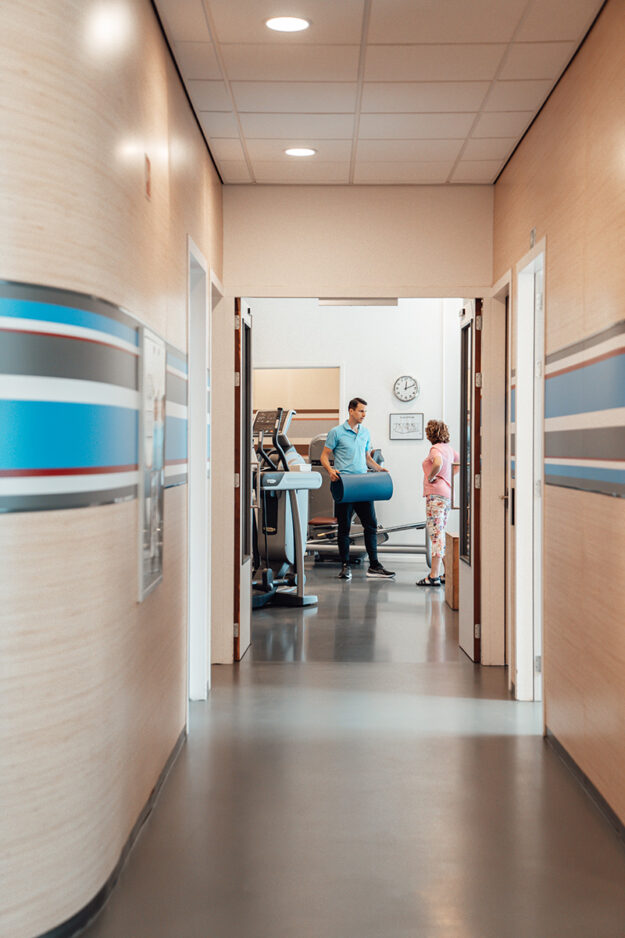 Aan het einde van de gang staat een fysiotherapeut met een patiënt in een oefenzaal. De patiënt deelt haar ervaring.