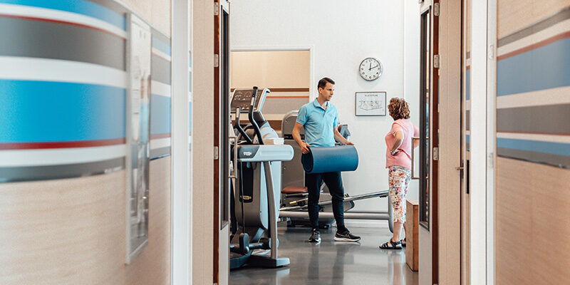 Ervaring Aan het einde van de gang staat een fysiotherapeut met een patiënt in een oefenzaal. De patiënt deelt haar ervaring.