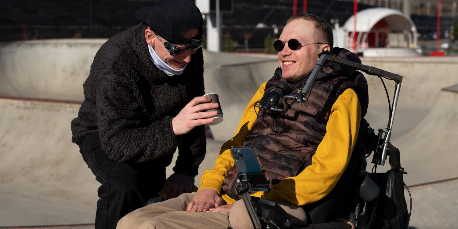 Man met ALS in een bodywarmer en gele mouwen wordt geholpen door een man in een zwarte jas