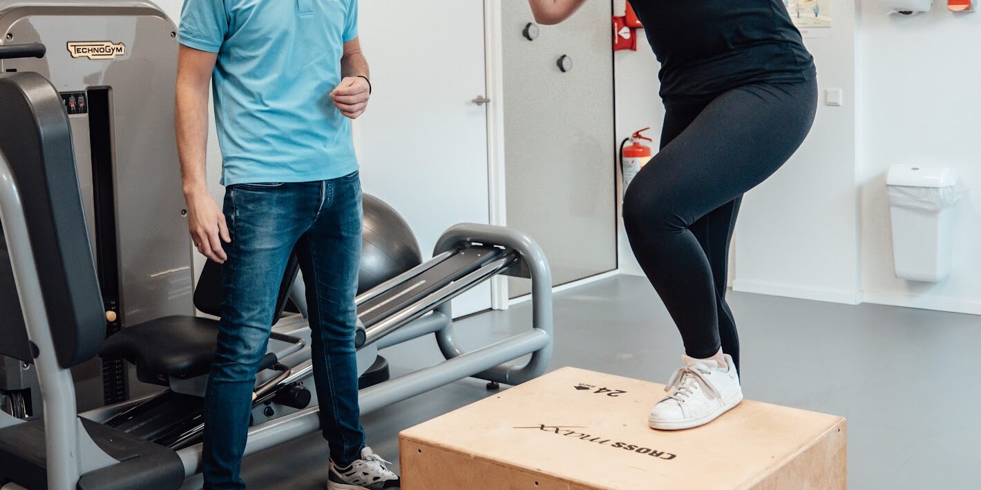 Vrouwelijke patiënt in zwarte kleding werkt weer aan de opbouw van haar spierkracht na het oplopen van een blessure met behulp van een fysiotherapeut in een blauwe polo
