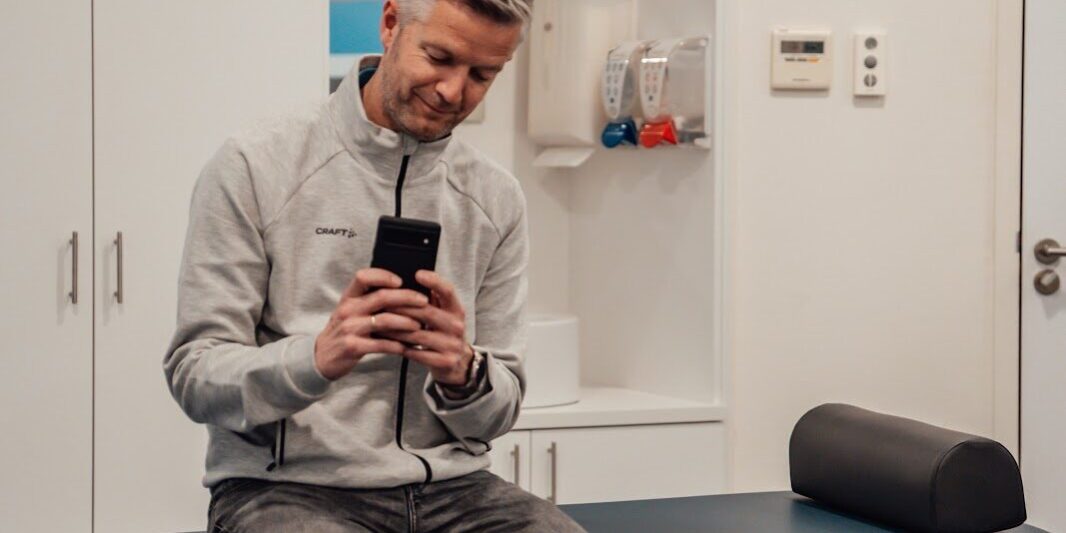 Social media Een man met een grijs vet op de behandeltafel van de fysiotherapeut met een telefoon in zijn hand op social media aan het scrollen