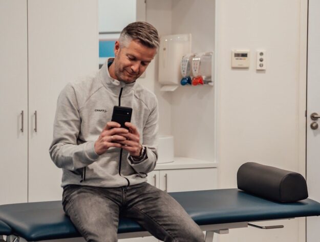 Een man met een grijs vet op de behandeltafel van de fysiotherapeut met een telefoon in zijn hand op social media aan het scrollen